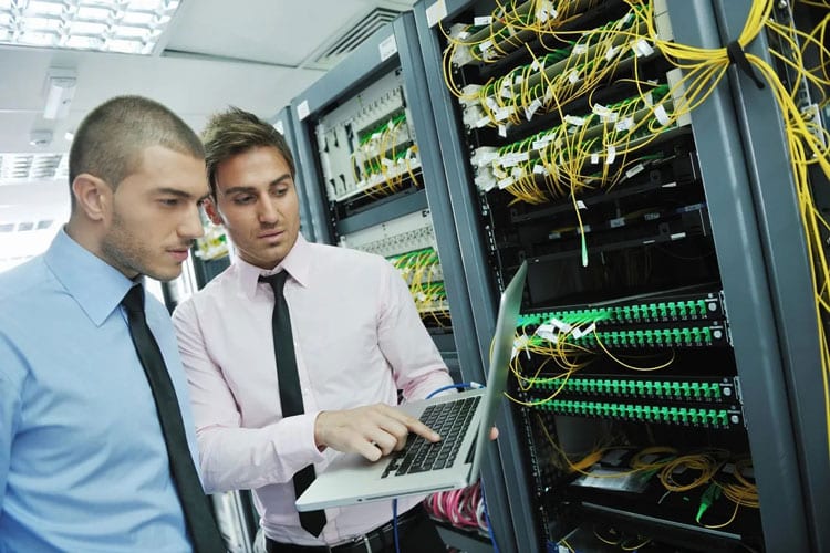Two men looking at a laptop in front of some servers.